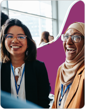 Two women wearing business attire and conference badges stand indoors at International House, smiling and talking; one wears glasses and a blazer, the other a headscarf and glasses.
