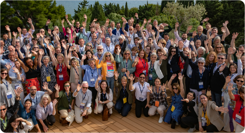Large group of people gathered outdoors at International House, smiling and waving at the camera, many wearing name badges and sunglasses, with trees and water in the background.