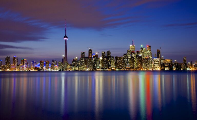 Toronto skyline at dusk with illuminated buildings and the CN Tower reflected on the calm water in the foreground.