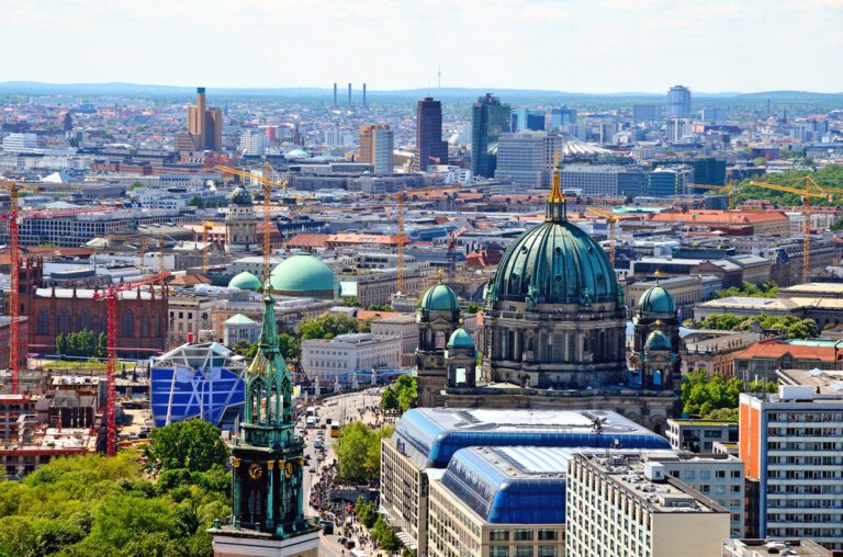 Aerial view of Berlin cityscape featuring the Berlin Cathedral, St. Mary's Church tower, modern buildings, and numerous construction cranes.