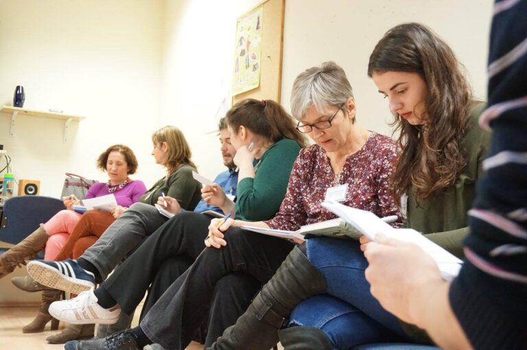 A group of adults sit in a row indoors, reading papers and writing notes during a classroom or workshop session.