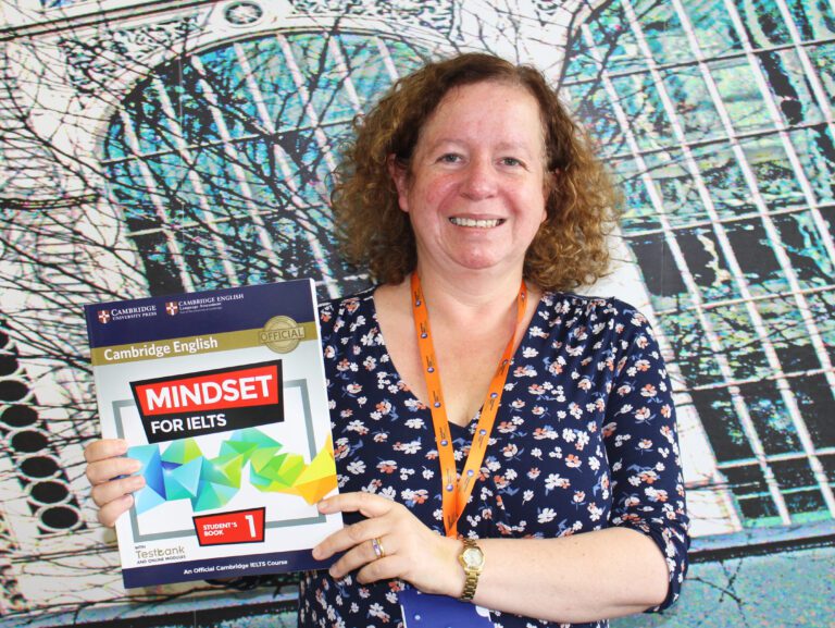 A woman holding a "Mindset for IELTS" Cambridge English textbook, smiling and standing in front of an abstract patterned background.