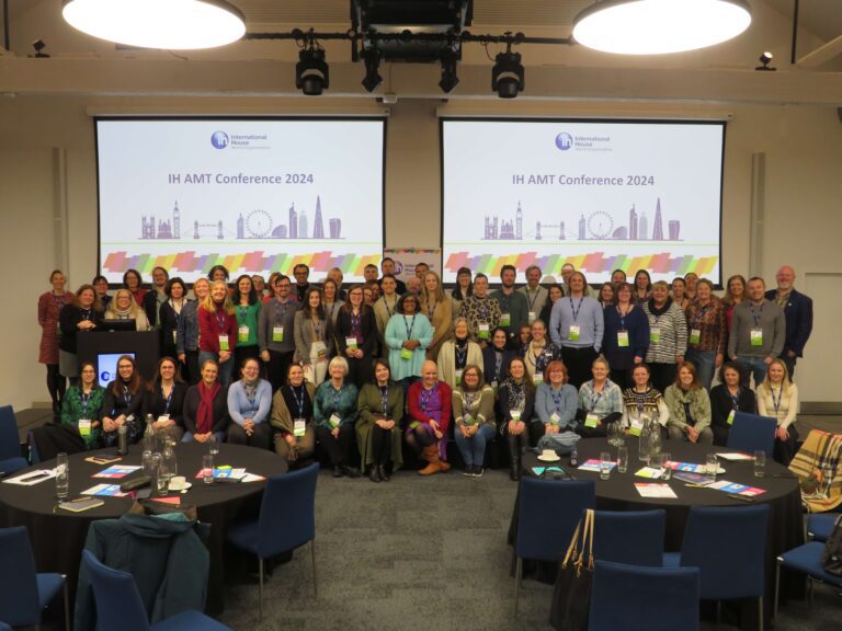 A large group of people pose together in a conference room with round tables; projected screens read "IH AMT Conference 2024.