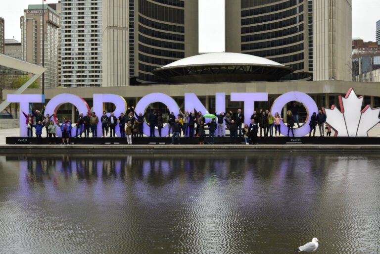 A group of people stand in front of a large illuminated "TORONTO" sign beside a reflective pool, with city buildings and a seagull in the foreground.