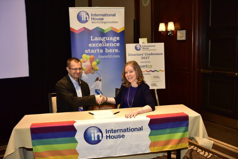Two people sit at a table covered with an International House banner, shaking hands in front of a conference sign that reads "Language excellence starts here.