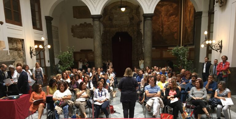 A woman stands speaking to a seated audience in a large hall with arched ceilings, columns, and decorative wall panels.