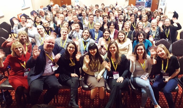 A large group of people seated in a conference room, many smiling and waving at the camera. Most are wearing name badges and lanyards.
