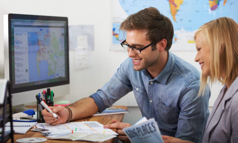 Two people sit at a desk with maps and a computer, smiling and planning a trip. World maps are displayed in the background.