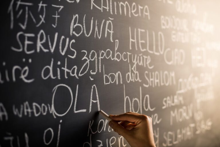 A hand writes greetings in various languages, such as "OLA" and "HELLO," on a blackboard with other multilingual phrases visible in chalk.