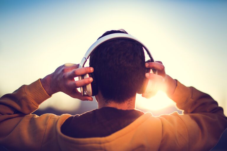 A person wearing headphones faces the sunrise or sunset, with their hands adjusting the headphones and light shining in the background.