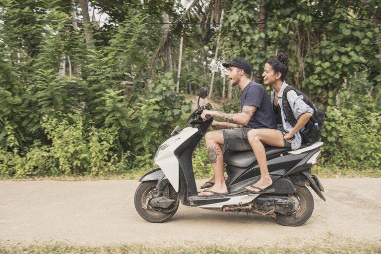 Two people ride a scooter on a rural road, surrounded by dense greenery. The driver wears a cap and shorts; the passenger carries a backpack and smiles.