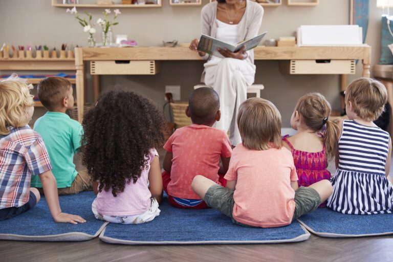 A group of young children sit on mats facing an adult who is reading a book in a classroom setting.