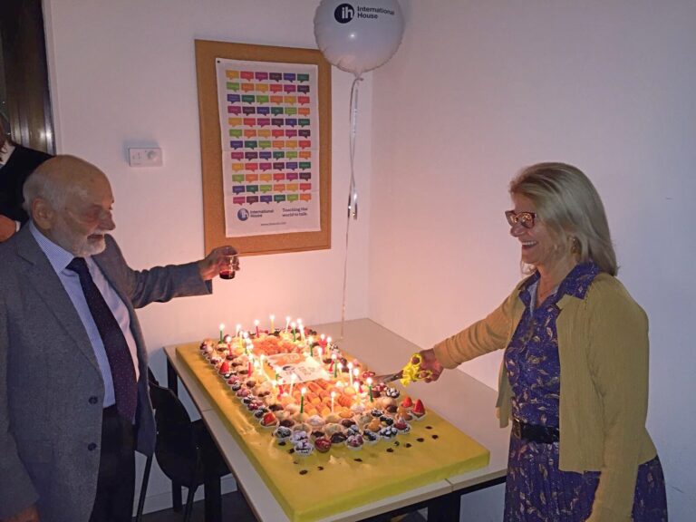 Two people stand by a table with a large, decorated cake topped with candles. The woman is cutting the cake while the man holds a glass. A balloon and colorful poster are in the background.