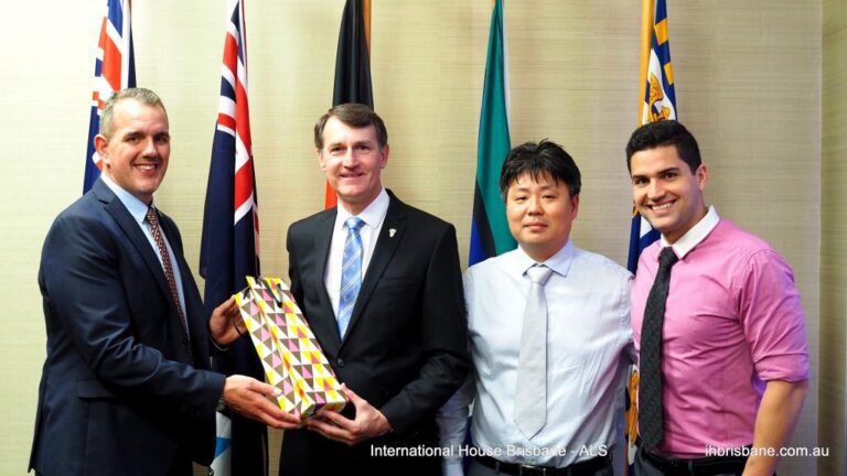 Four men in business attire stand in front of several flags; one man hands a patterned gift box to another.