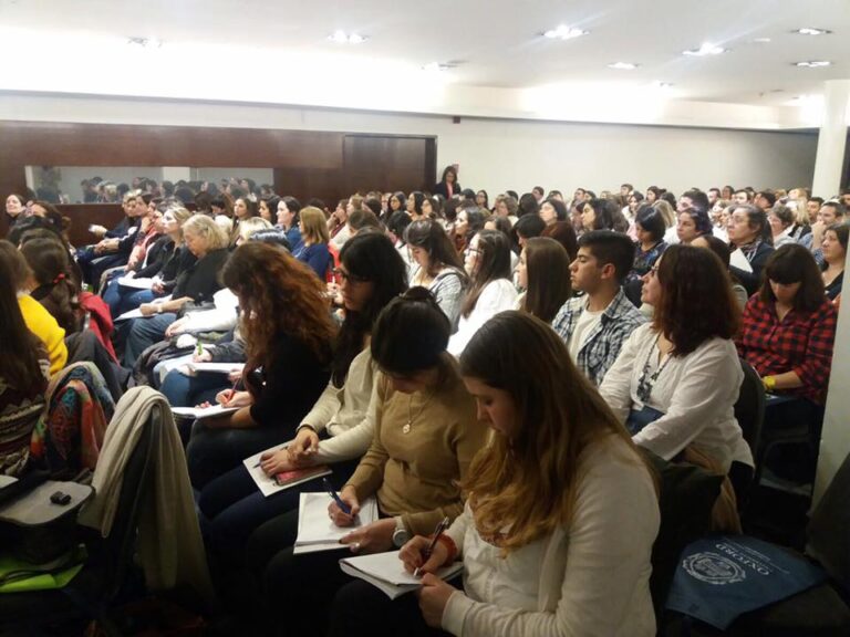 A large group of people sits in rows inside a conference room, many taking notes and listening attentively to a speaker out of the frame.