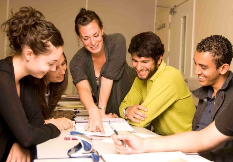 A group of five young adults sit around a table with notebooks, smiling and discussing as one person points to something in a book.