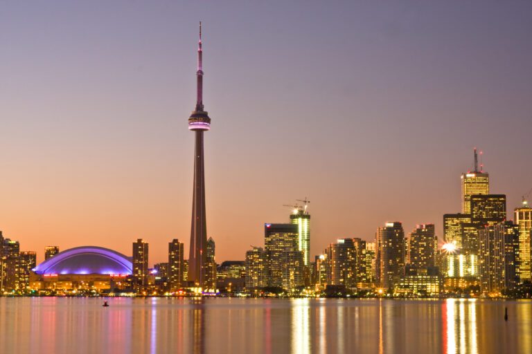 Toronto skyline at dusk, featuring the CN Tower, Rogers Centre with a purple dome, and city buildings reflected in calm water.