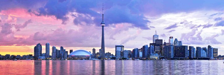 Toronto skyline at sunset, with the CN Tower and Rogers Centre visible, reflected in the water under a partly cloudy sky.