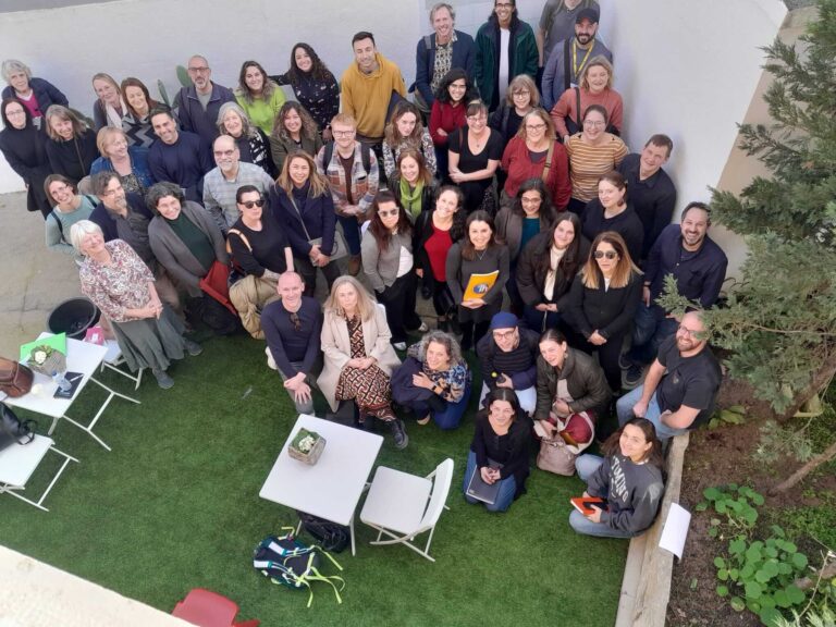 A large group of people pose for a photo outdoors in a courtyard with artificial grass, surrounded by white walls and a few tables and chairs.