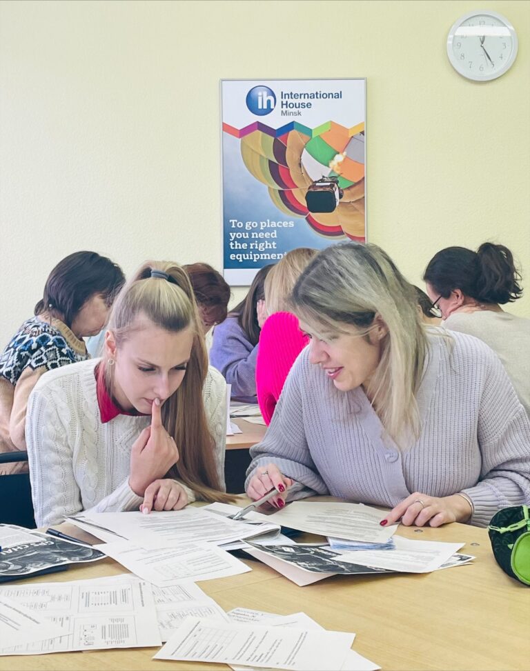 Two women sit at a desk, examining papers together in a classroom setting, with other people working in the background and a poster and clock on the wall.