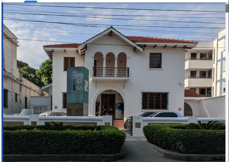 A two-story white building with a red-tiled roof, arched entrance, and glass sign in front, surrounded by a low white fence and parked cars.