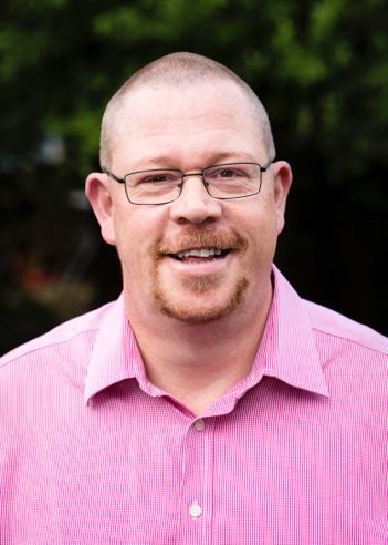 Man with short hair, glasses, and goatee smiling, wearing a pink collared shirt, with a blurred green background.