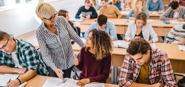 A teacher stands beside a student, helping her with classwork, while other students work at desks in a bright classroom.