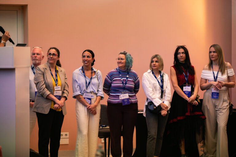 Seven people wearing conference badges stand in a row facing the audience inside a room with peach-colored walls, representing our About Us team.