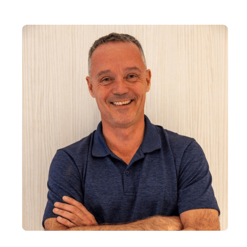 A man with short gray hair wearing a navy blue polo shirt stands in front of a light-colored wooden wall, smiling with his arms crossed, reflecting a celebration of diversity through his welcoming and confident presence.