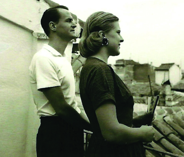 A man and a woman stand side by side on a rooftop terrace, looking out over buildings on a cloudy day. This black and white image captures an authentic moment that reflects the spirit of our About Us story.