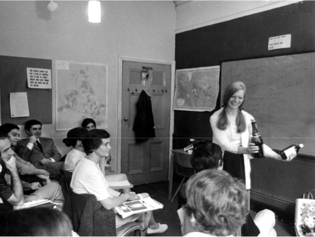 A woman stands at the front of a classroom holding two bottles while several students seated at desks watch her. Maps and notes, including an About Us poster, are visible on the walls.