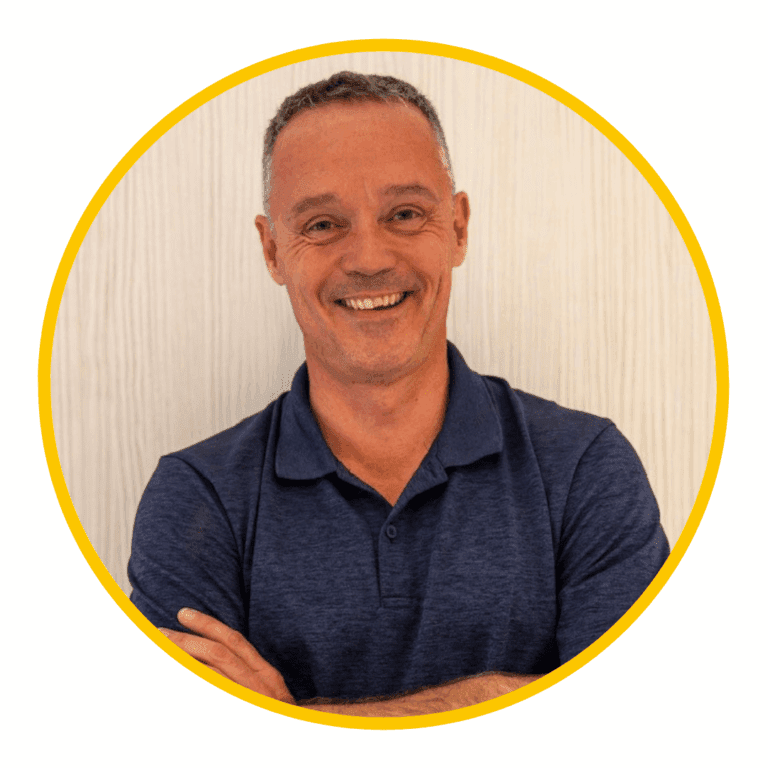 Smiling man with short gray hair, wearing a navy blue polo shirt, stands with arms crossed in front of a light background framed by a yellow circle—reflecting a Celebration of Diversity.