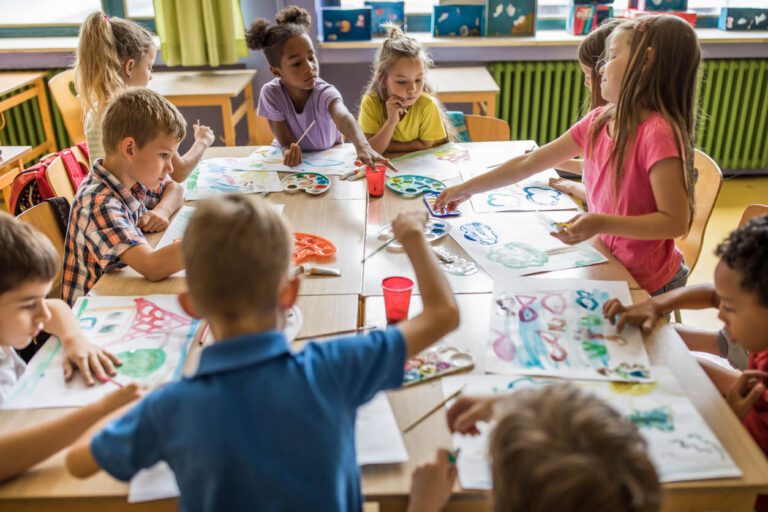 A group of young children sit around tables in a classroom, painting and drawing with art supplies on sheets of paper.