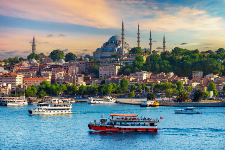 Boats travel across the Bosphorus with a cityscape view of Istanbul, featuring the prominent domes and minarets of a large mosque under a colorful sky.