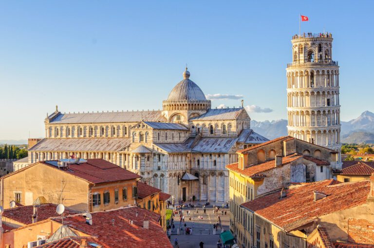 The Leaning Tower of Pisa stands next to Pisa Cathedral, surrounded by red-roofed buildings under a clear sky.