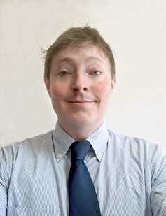 A person with short light brown hair wearing a light blue striped shirt and a navy blue tie, smiling at the camera against a plain light background.