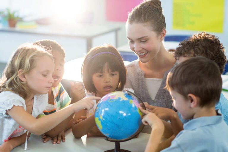 A teacher and five young children look at and point to a globe in a classroom.