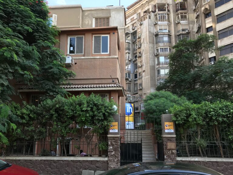 A two-story tan building with barred windows and a green hedge in front, surrounded by taller apartment buildings and trees.
