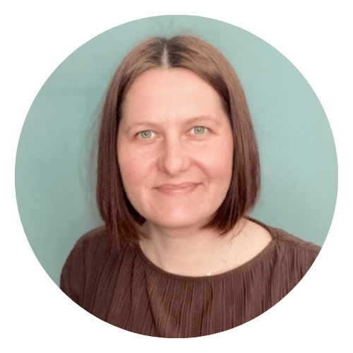 A woman from the IHWO Team with straight, shoulder-length brown hair and a brown top smiles at the camera against a plain light blue background.