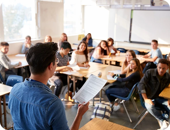 A teacher stands at the front of a classroom, holding papers and addressing students who are seated at desks and listening attentively.