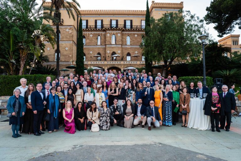 A large group of International House Directors in formal attire pose for a group photo outdoors in front of a historic multi-story building with palm trees and greenery.