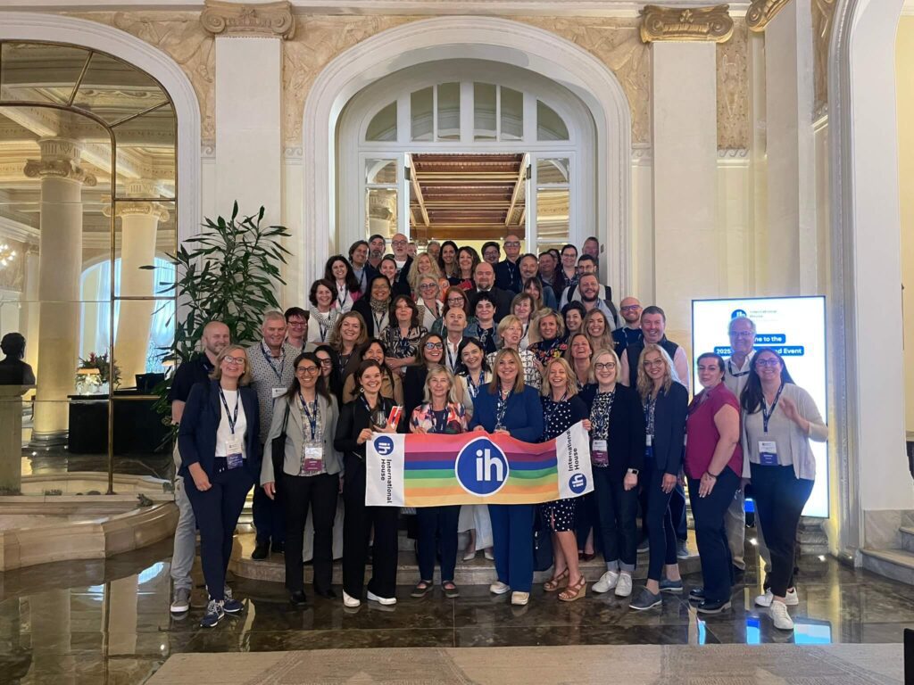 A large group of people poses indoors on a staircase, holding a rainbow-colored "IH" banner; most are wearing ID badges and business attire.