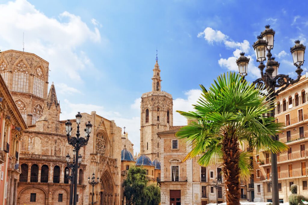 A historic city square with Gothic and Baroque buildings, including a cathedral, a bell tower, palm trees, and ornate street lamps under a partly cloudy sky.