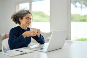 Woman with curly gray hair and glasses sits at a desk holding a mug, looking at a laptop screen, with a notebook and pen nearby in a bright room. She is studying an International House Online Teacher Training Institute course.