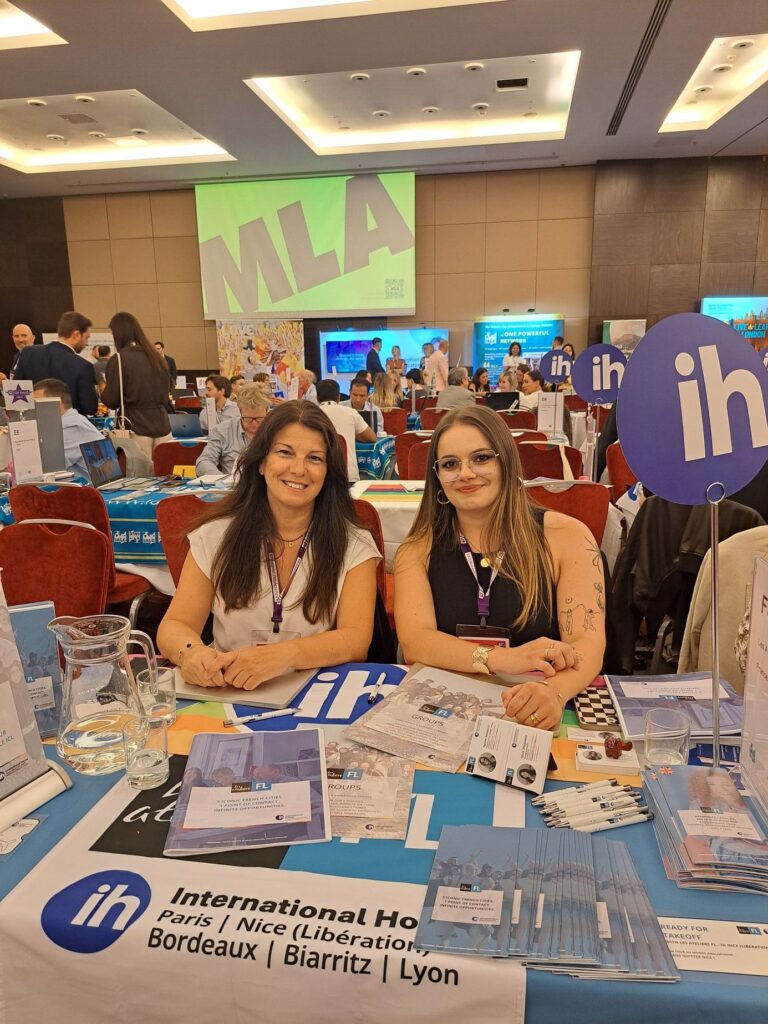 Two IH members smile whilst sat behind a table decorated with ih banner and ih globe on a pole. The banner on the table reads 'International House Paris | Nice (Libération) | Bordeaux | Biarritz | Lyon'