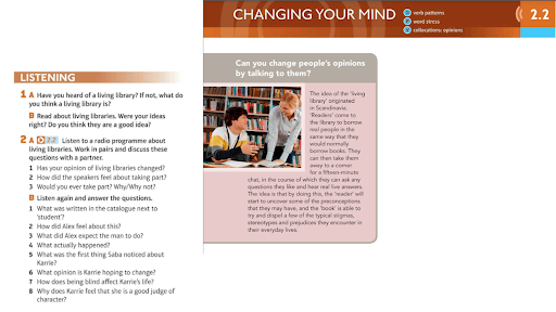 Two students sit at a table in a library, talking and looking at books. A third person stands in the background near shelves filled with books.