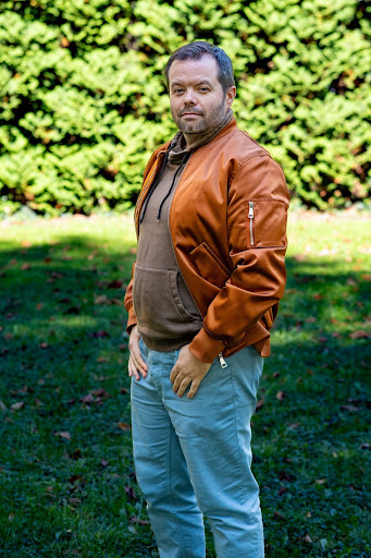 A man in a brown leather jacket poses side-on in a garden in front of a hedge.