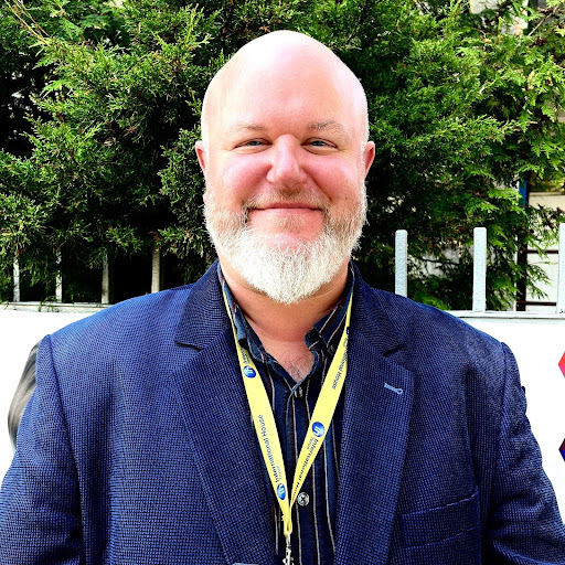 A bald man with a white beard and navy suit jacket smiles at the camera standing in front of fir trees.
