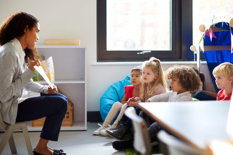 A woman teacher sits in front of a group of children showing them a book.