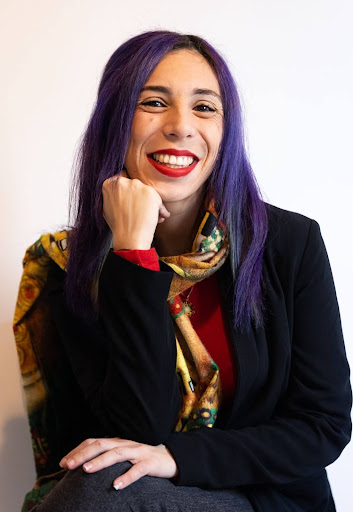 A woman with purple hair and red lipstick rests her head on her hand and crosses legs smiling widely at the camera.